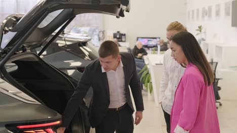 young car salesman showing to young couple new automobile at dealership salon.