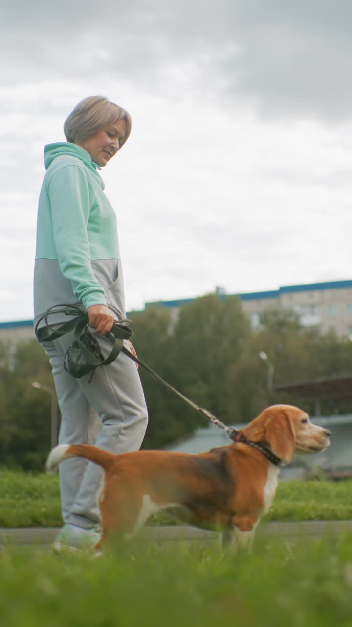 코카서스인 트레이너가 비글을 데리고 길을 따라 활기차게 걷고 있어요. 운동화와 후드티를 입고 공원과 들판 근처에서 끈을 잡고 집중하며 걷고 있으며, 하늘은 흐린 날씨입니다. 활기찬 낮 산책과 기본적인 복종 훈련을 하고 있어요.