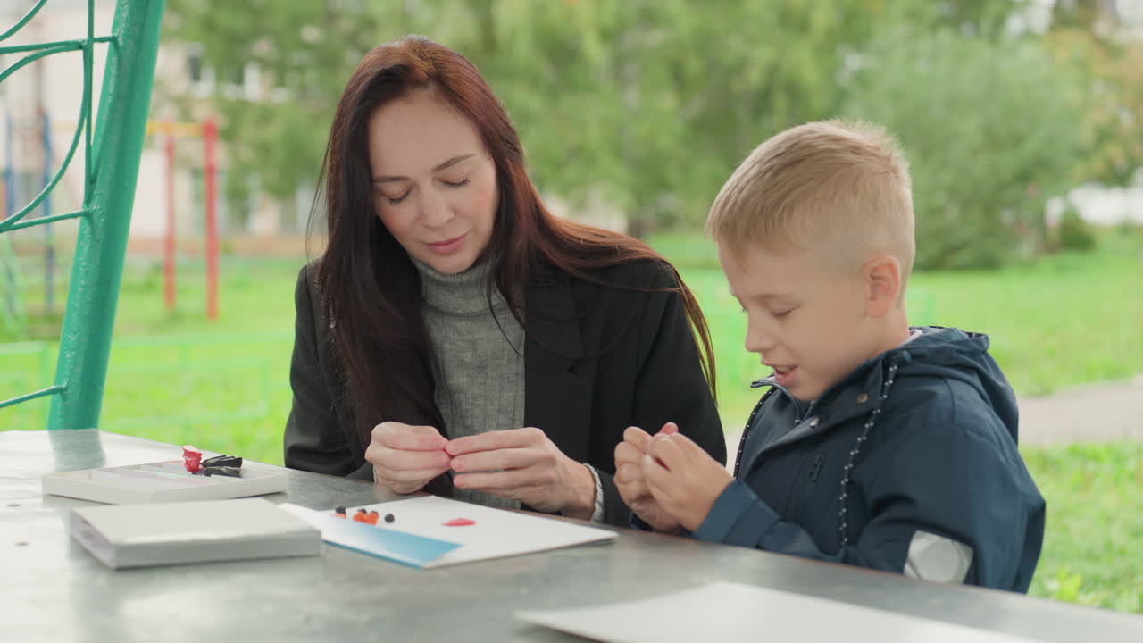Hvit mentor forbereder skolemateriell utendørs ved et piknikbord, ung sønn viser frem et håndverk mens en moderlig veileder tilbyr mild veiledning og oppmuntring, praktisk aktivitet, grønn parkbakgrunn.