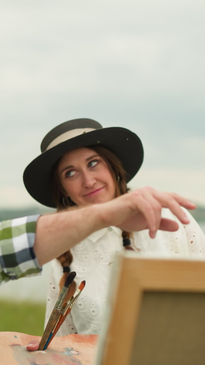 A man in a black hat and plaid shirt stands close to a woman in a white dress in a scenic field. He gestures in a direction, possibly discussing their next steps or an art composition