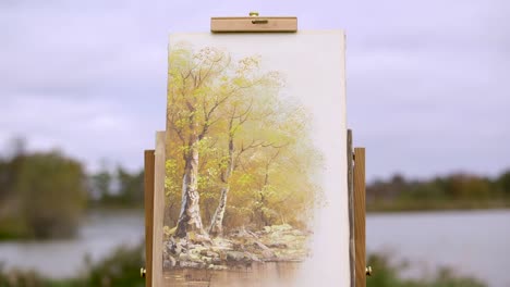 painting of trees sits outside in front of a lake out of focus