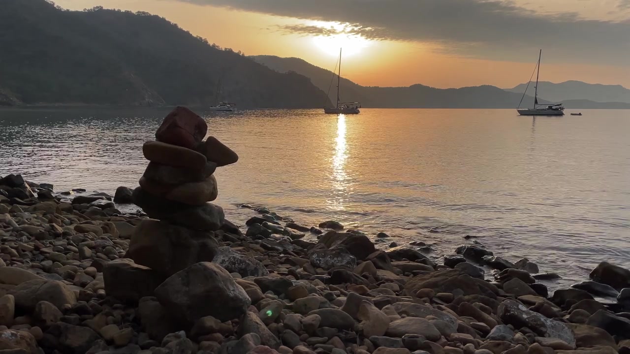 Turquía, arte de equilibrio de rocas en una playa desierta de guijarros. La cámara enfoca el equilibrio de rocas y los dos barcos anclados en una bahía tranquila durante el atardecer.