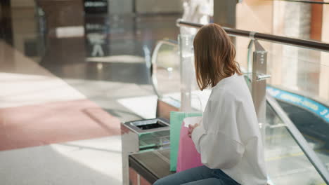 woman seated in a shopping mall retrieving a yellow-covered book from a pink bag, with a blurred escalator in the background and a passerby approaching the moving escalator