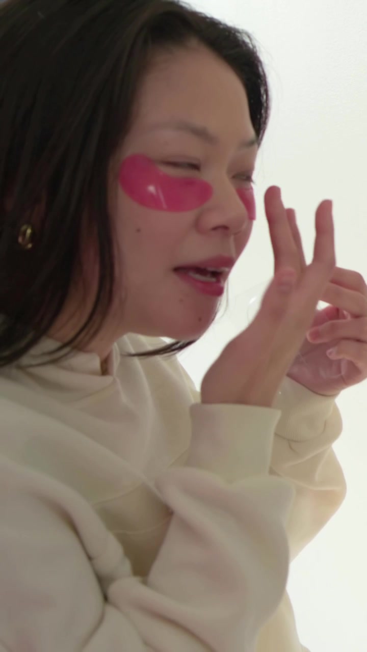Vertical video: Inspecting compact, woman applying pink under-eye patches for care in bright room