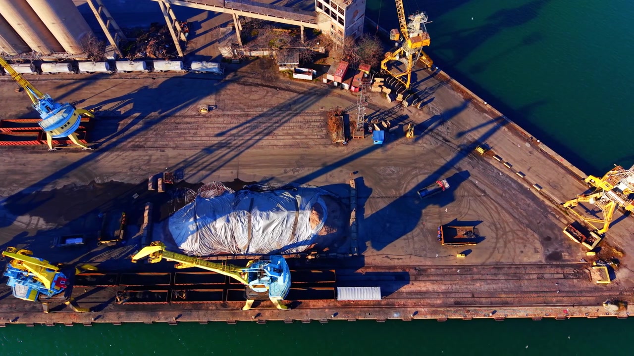 Overhead view of a dock crane and cargo pile. Yellow machine casting a long shadow on concrete ground next to a covered material heap.