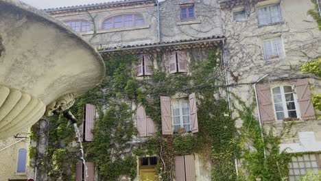 Fountain-in-front-of-a-beautiful-old-French-house-with-lots-of-hanging-plants-made-of-stone-with-railed-windows-and-shutters