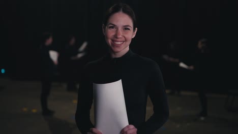 Portrait-of-a-happy-and-confident-young-brunette-girl-in-a-black-actress-costume-who-holds-sheets-of-script-and-character-text-in-her-hands-during-her-preparation-for-a-performance-in-the-theater