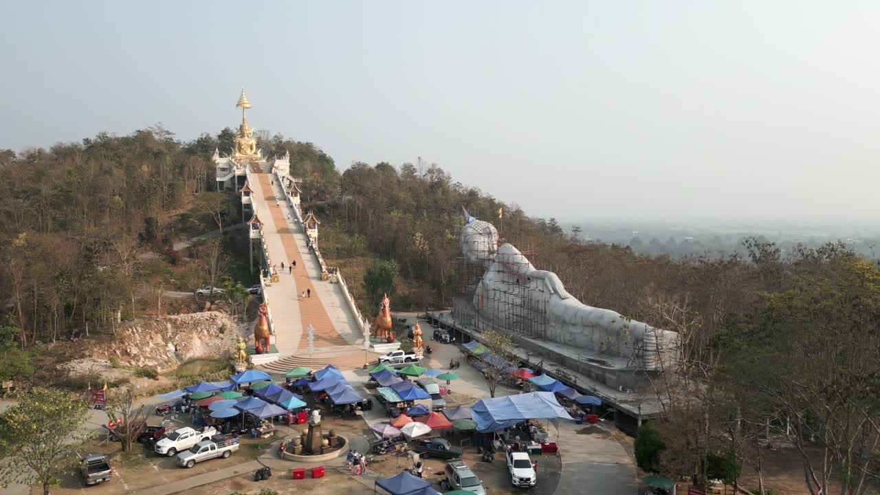 Aerial view of a large reclining Buddha statue under construction, a temple with a golden Buddha at the top of a long staircase, and a market below
