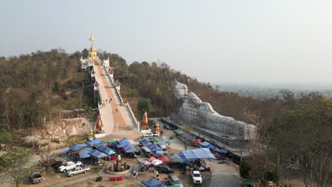 aerial view of a large reclining buddha statue under construction, a temple with a golden buddha at the top of a long staircase, and a market below