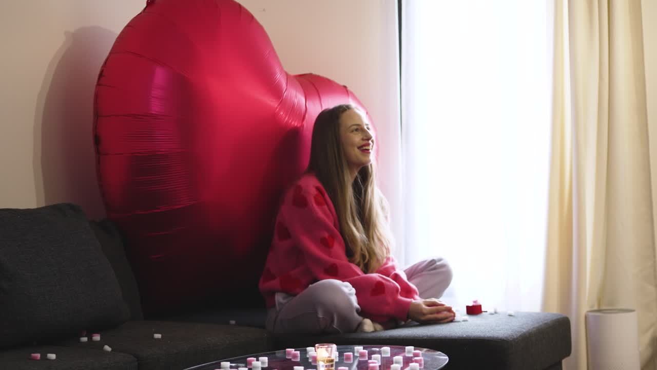 Una giovane donna seduta su un divano con grandi palloncini a forma di cuore dietro di sé, lancia piccoli cuori in aria, ballando e festeggiando San Valentino con gioia in un accogliente salotto