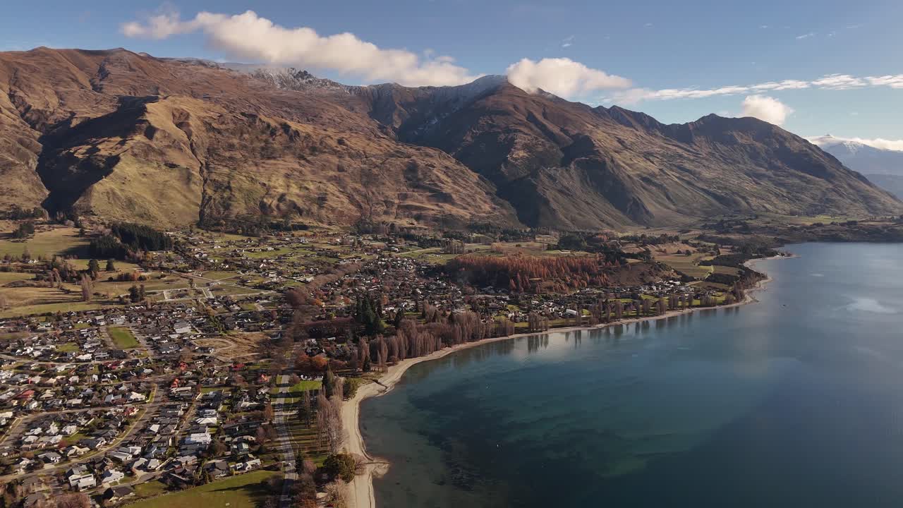 Wanaka town with beautiful coastline during autumn season. Sunny day In New Zealand. Aerial wide shot. Tranquil lake, houses along shoreline and leafless trees at beach. Mountains in back