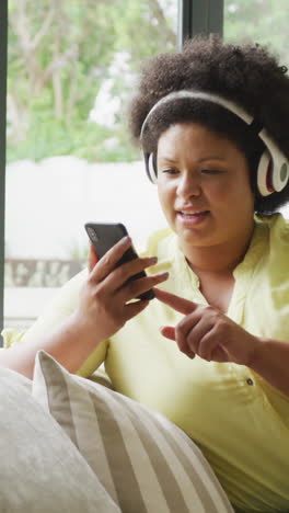 video of happy plus size african american woman with headphones sitting on sofa with smartphone