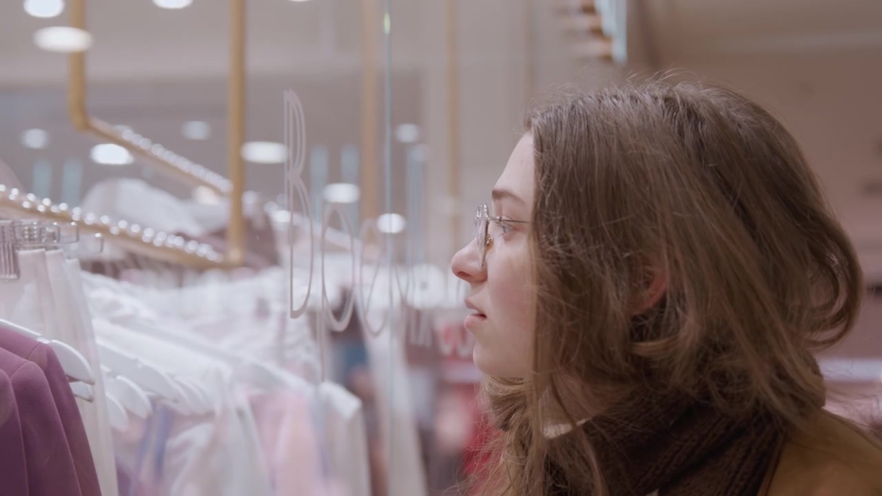Caucasian woman browsing boutique clothing racks, studying hangers of sweaters and dresses with thoughtful expression glasses and coat visible, muted mall lights and neat displays set calm, stylish