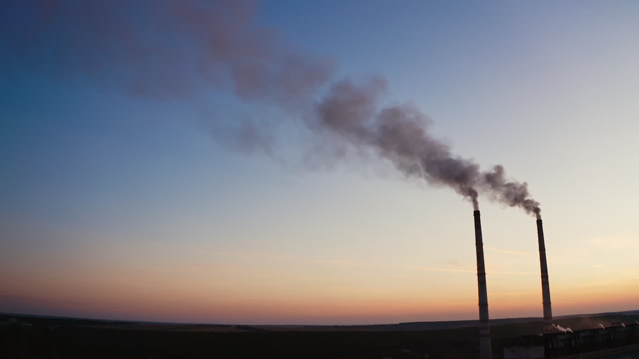 Pipes with smoke on the evening sky background. Harmful smoke going into the air from two industrial pipes of a factory at sunset.