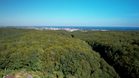 Drone-forward-flight-over-forest-landscape-with-Gdynia-City-and-Sea-in-background-during-sunny-day-with-blue-sky,-Poland