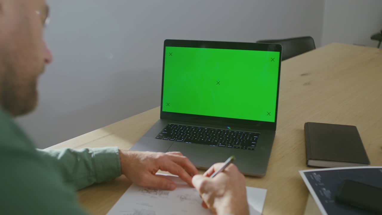 un hombre está trabajando en un dibujo, sentado frente a una computadora portátil con una pantalla verde