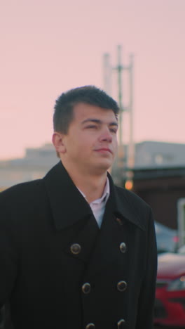 young man in black coat exits car holding briefcase in left hand, background features parked cars, office residential building, and red cloud in sky