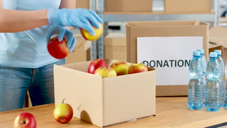 Vista-Cercana-De-La-Caja-De-Embalaje-De-Manos-Voluntarias-Con-Manzanas-Rojas-Y-Agua-En-El-Almacén-De-Caridad