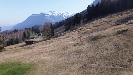 Drone-fly-along-dry-field-of-grass-in-winter-with-small-huts-and-snowy-mountains-in-background