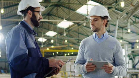 Kaukasischer-Männlicher-Arbeiter,-Der-Einen-Helm-Trägt-Und-Ein-Tablet-In-Einer-Fabrik-Benutzt,-Dann-Kommt-Sein-Kollege-Und-Spricht-Mit-Ihm