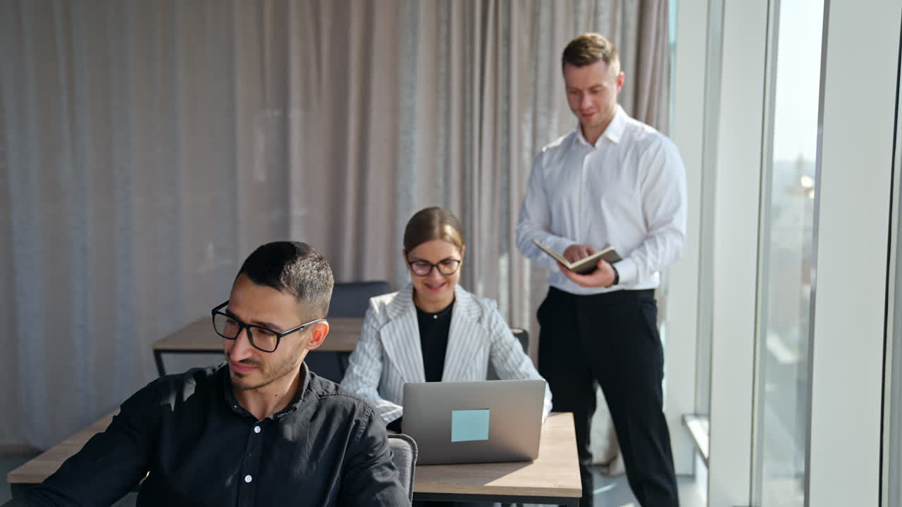 Male manager checking information in his paper notebook standing near female employee. People using computers at their work in office.