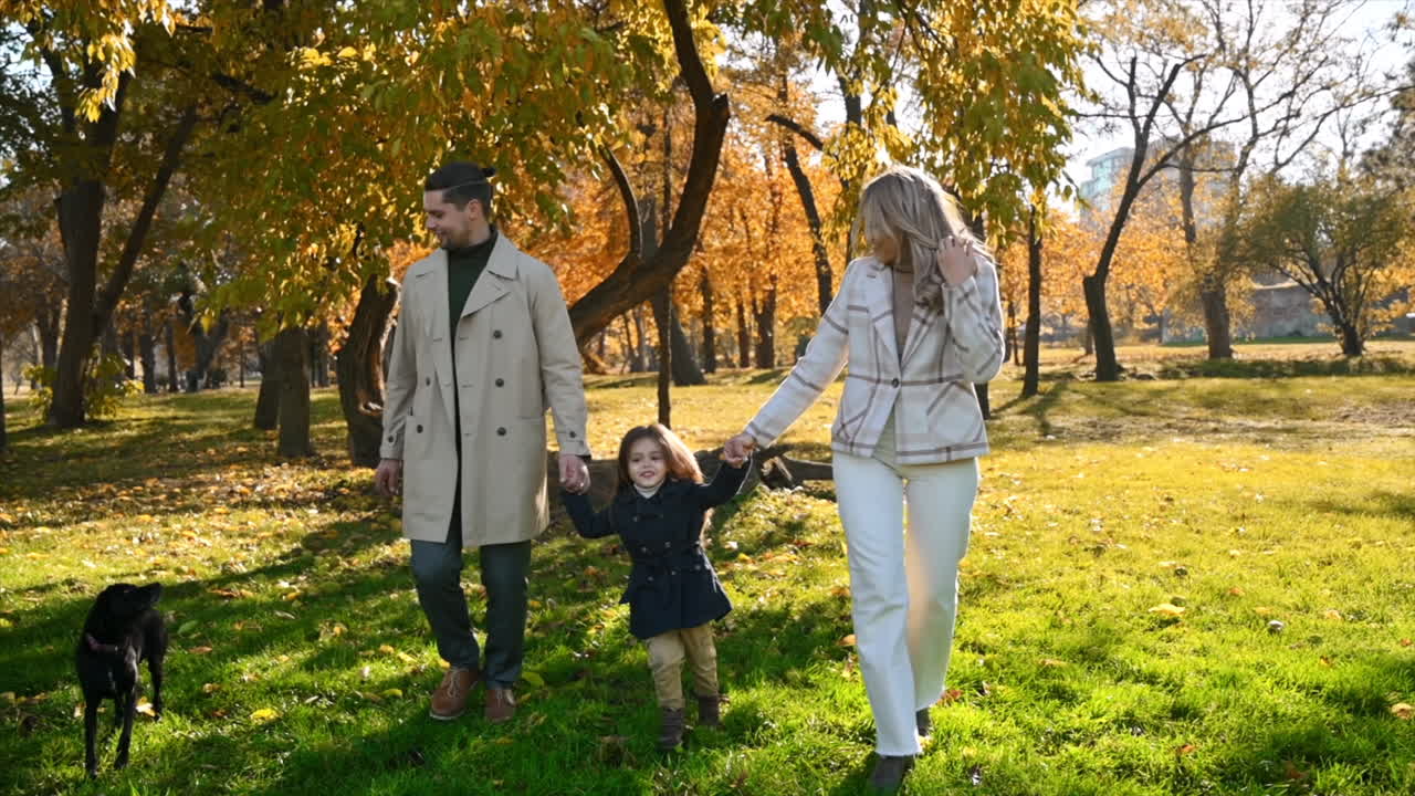 Happy family in an autumn park. Mother and father slowly running with daughter and their dog, yellowed trees around. Slow motion