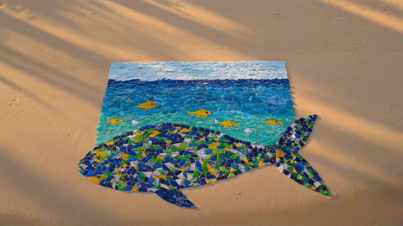 Un primo piano sulla spiaggia mostra le mani di un bambino che posizionano frammenti traslucidi di vetro marino in un motivo a pesce e onda sulla sabbia umida, con una luce dorata soffusa, texture e una giocosa creatività artigianale.