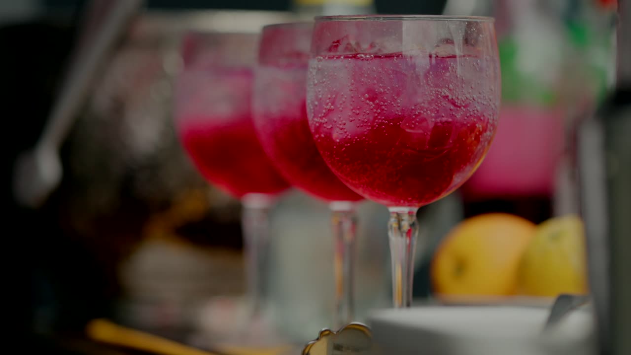 Three glasses of red cocktails with ice, lined up on a bar with fruits blurred in the background.