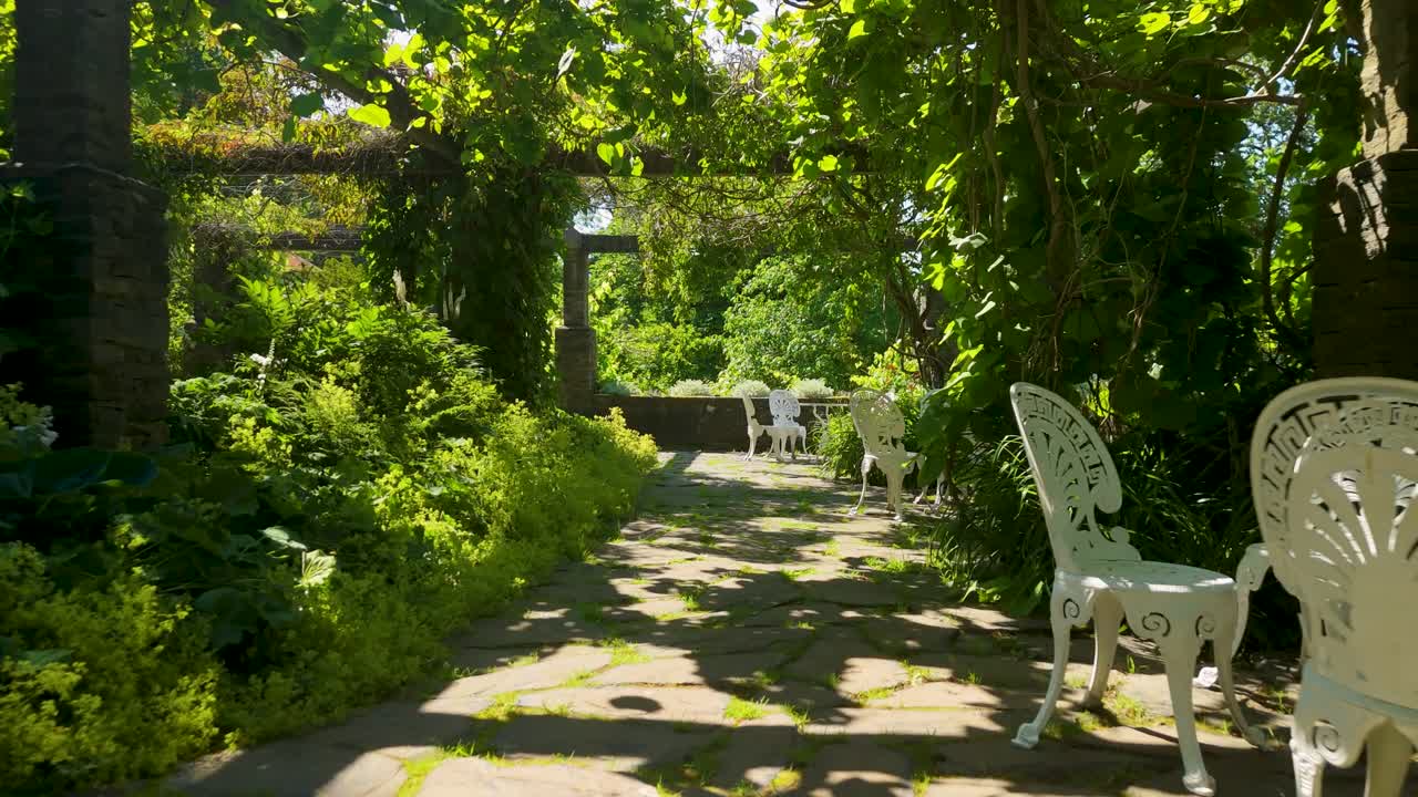 La cámara avanza a través de un exuberante jardín verde hacia una zona de asientos con pérgola pavimentada en piedra, con muebles de hierro forjado blanco y plantas trepadoras en la casa señorial Baldersnäs Herrgård en Dalsland, Suecia.