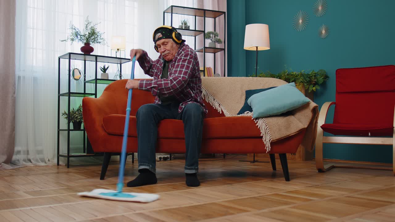 Happy senior grandfather housekeeper is mopping floor in living home room listening music dancing