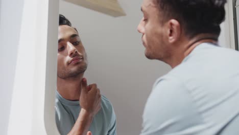 focused biracial man looking in sunny bathroom mirror, examining his face and skin, slow motion