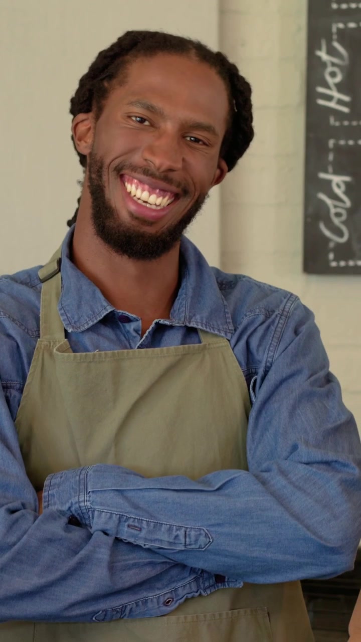 Vídeo vertical: Ouvindo a deixa, funcionário de cafeteria rolando tablet atrás do balcão, com avental verde-oliva, sorrindo.