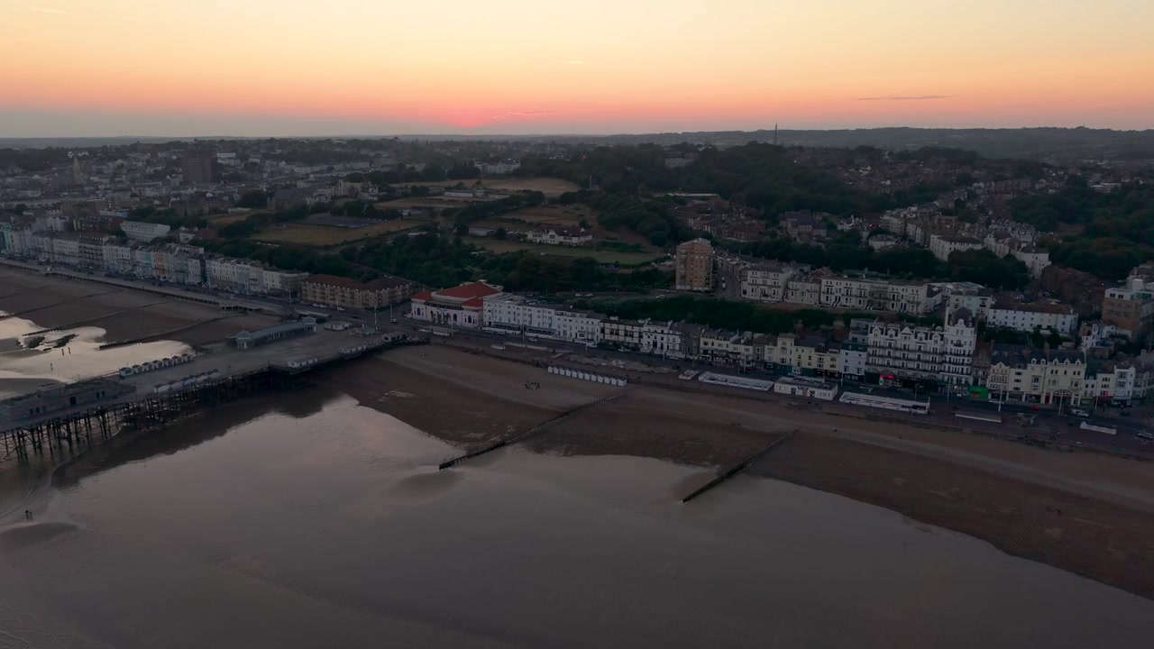 Drone vola veloce sul mare verso Hastings Pier, superando il White Rock Theatre e il lungomare al tramonto, East Sussex, Inghilterra
