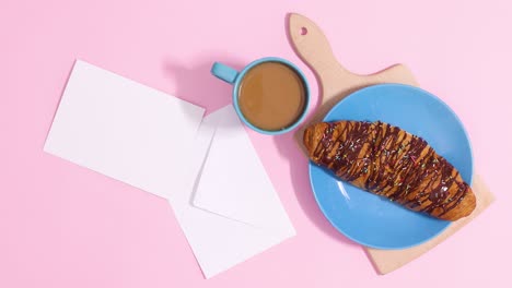 el croissant de chocolate aparece con una taza de café caliente y una nota de tarjeta de papel con sobre sobre un fondo rosa pastel.