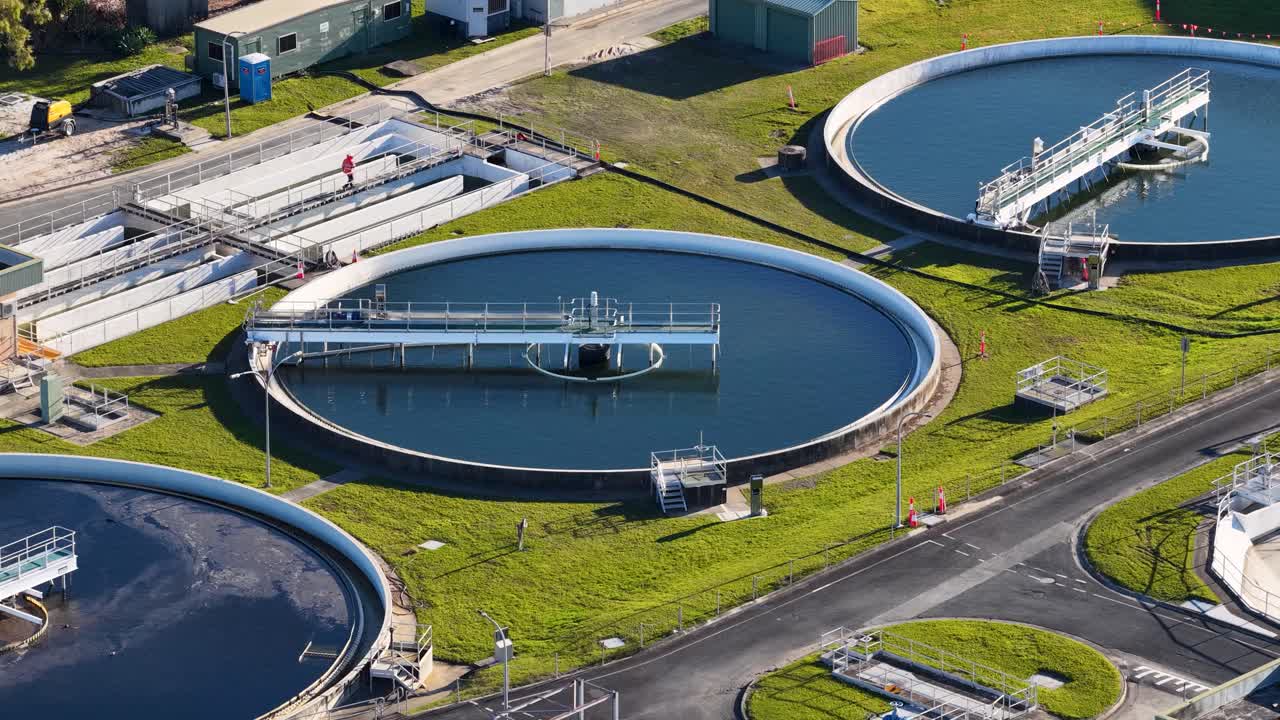 Rotating camera captures circular water treatment tanks, industrial structures, and a worker at a modern sewage facility under bright daylight in Gold Coast, Australia