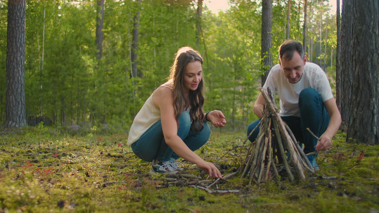 улыбающаяся женщина и мужчина вместе в лесу собирают и устанавливают палки для костра на закате во время семейного кемпинга