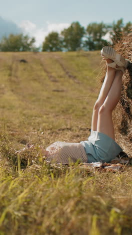 dama en cuclillas tomando fotos de su hermana al aire libre mientras coloca las piernas en el heno en vastas tierras de cultivo, cálido entorno rural con campos dorados, atmósfera rústica y luz solar que arroja sombras suaves
