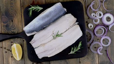 fillet of salted herring with onions, lemon and rosemary on dark cutting board rotating.