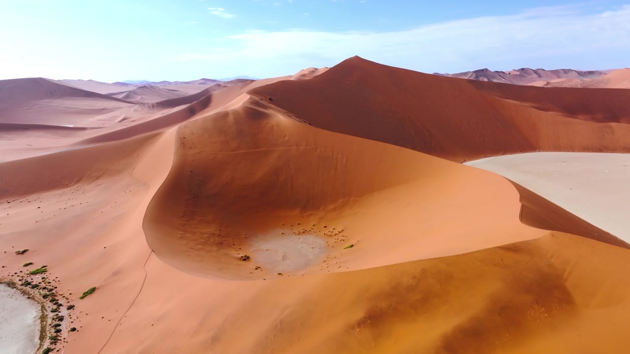 Vista de drone de uma duna vermelha esculpida encontrando a planície de argila branca de Sossusvlei sob céu azul. As imponentes dunas vermelhas brilham contra as planícies de argila pálidas e rachadas desta antiga paisagem desértica selvagem