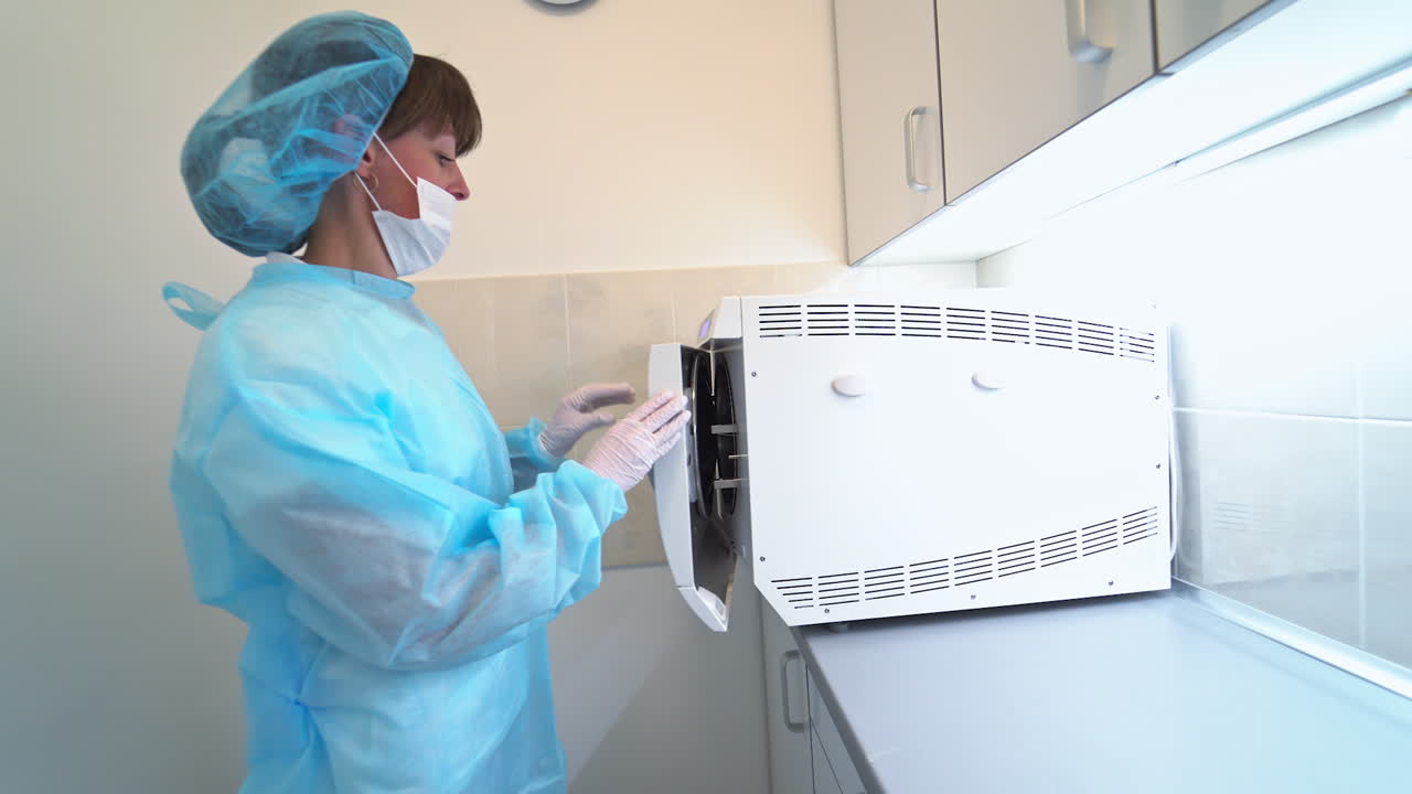 Female medical care professional in protective clothes and mask. Dental nurse puts some dental care instruments into sterilizer equipment. Side view.
