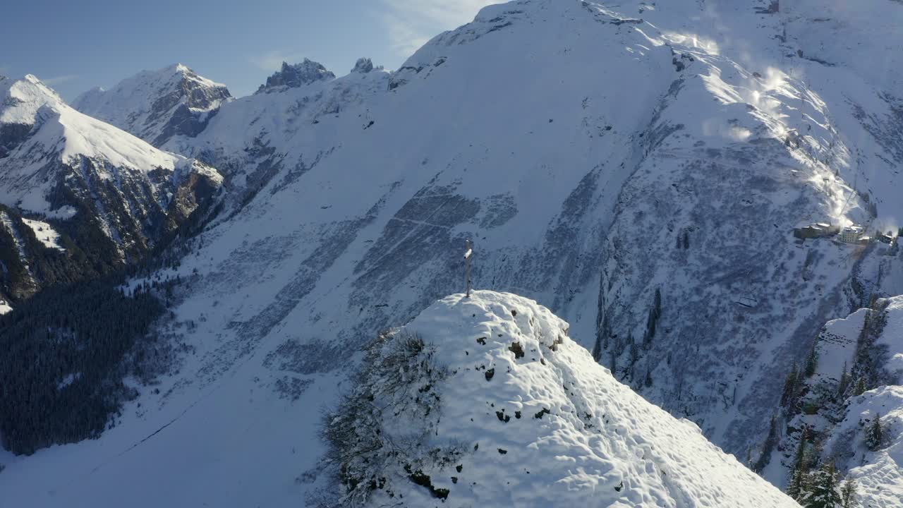 Aerial footage of slow orbit around mountain top with a summit cross covered in fresh snow, with the town of Engelberg in the background