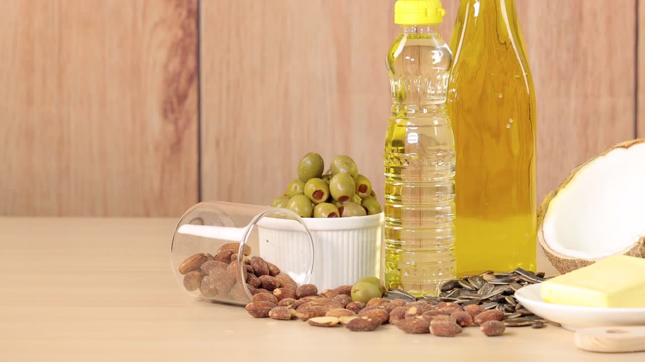 A variety of high-fat foods including avocado, coconut, bacon, butter, nuts, oil, and olives arranged on a wooden table, captured in bright studio lighting with a smooth left-to-right pan