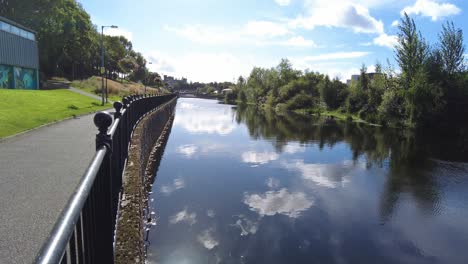 Tiro-Ascendente-Que-Muestra-La-Pasarela-A-Lo-Largo-Del-Río-Nore-En-Kilkenny-Irlanda-Con-El-Castillo-De-Kilkenny-En-La-Distancia