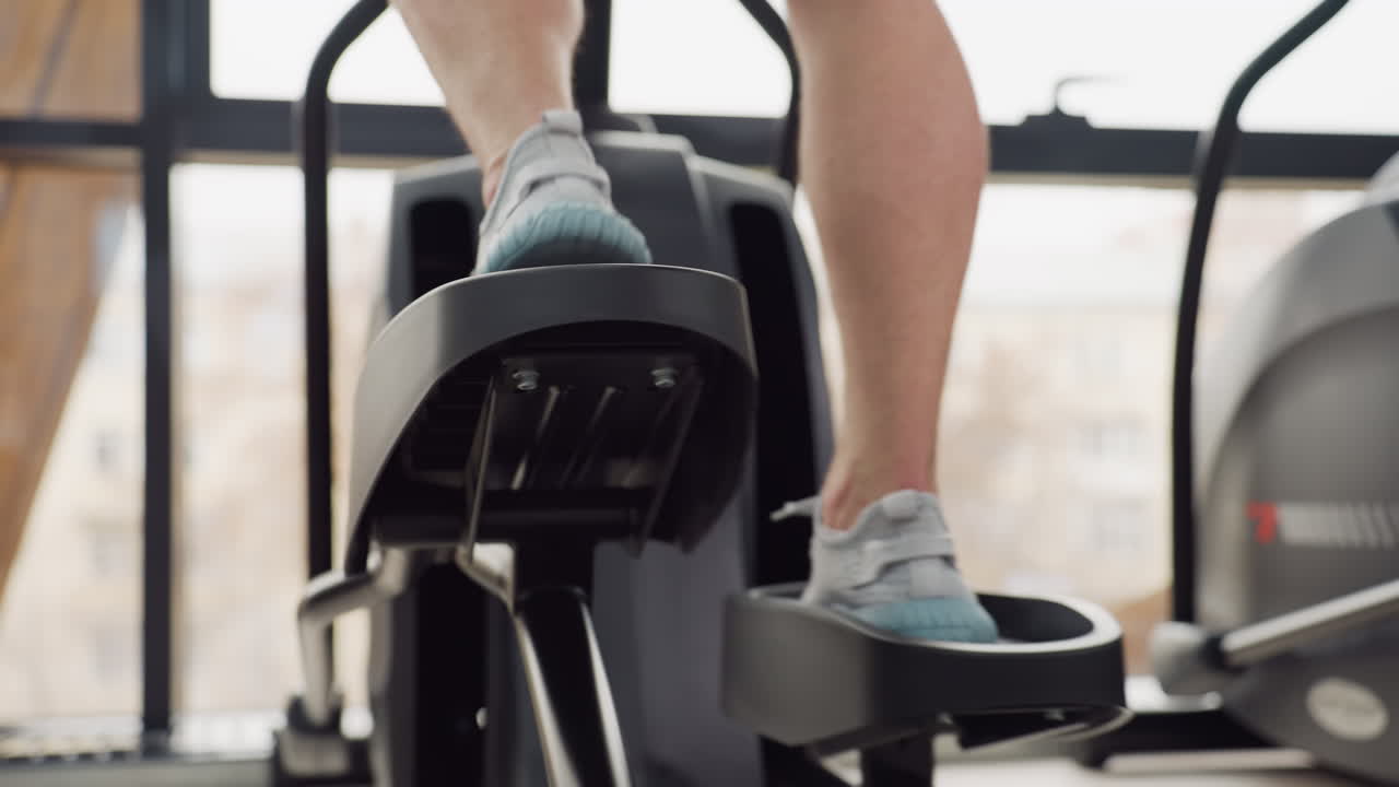 Close up of gym enthusiast wearing sneakers powering elliptical trainer pedal with controlled motion during dynamic cardio workout in bright sleek modern fitness center by large windows