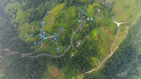 Panoramic-drone-top-down-overview-of-rural-farmland-village-with-terraced-fields-and-bright-blue-roofs,-misty-cloud-haze