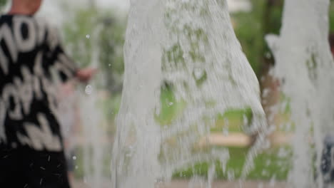 young girl near water fountain in park while mother walks toward her, splash from fountain in foreground, green grass and trees blurred background, summer afternoon family outing