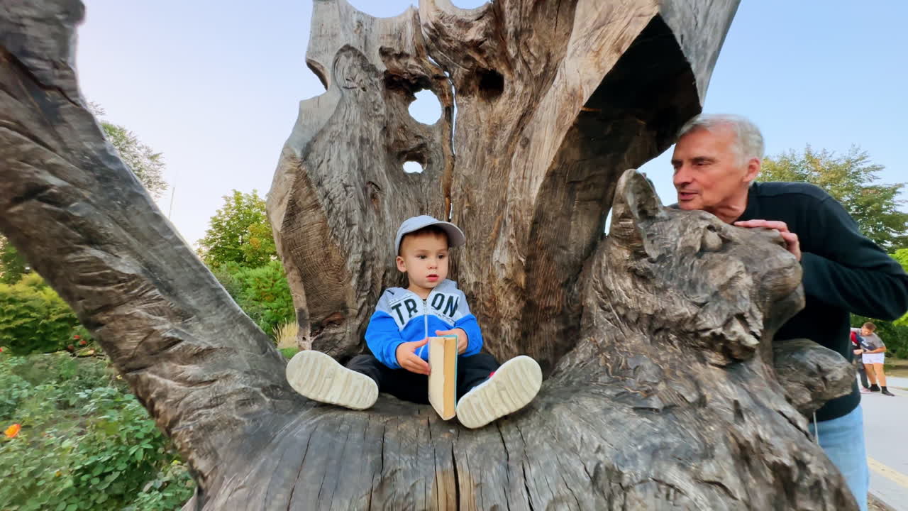 Lovely baby boy in cap sits in the chair carved in the wood holding a book. Grandpa stands beside talking to a grandson.