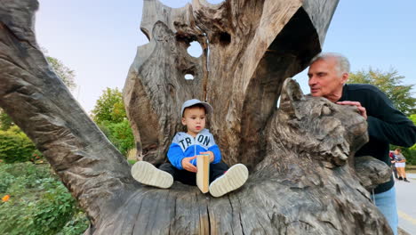 lovely baby boy in cap sits in the chair carved in the wood holding a book. grandpa stands beside talking to a grandson.