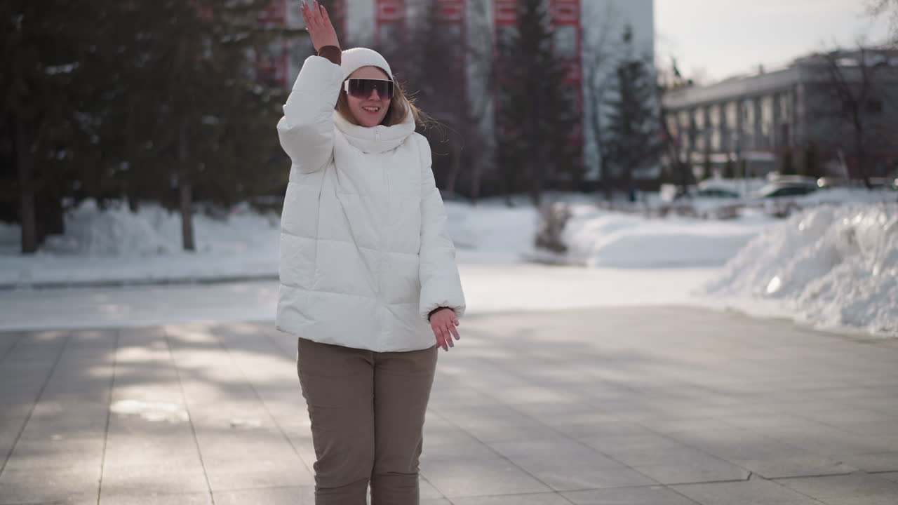 Close up of energetic young woman in white winter coat, beanie and sunglasses performing hip hop dance moves on snow covered street pavement while pedestrian crosses behind under bright daylight