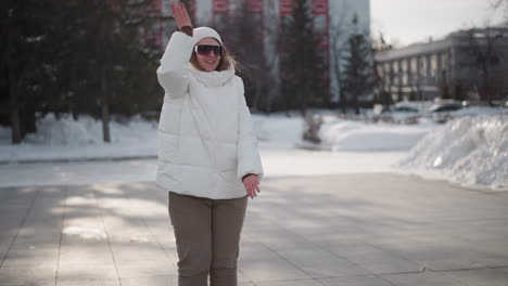 close up of energetic young woman in white winter coat, beanie and sunglasses performing hip hop dance moves on snow covered street pavement while pedestrian crosses behind under bright daylight